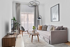 A modern living room with a grey sofa, a round wooden coffee table, a black floor lamp, and a large window with sheer curtains. - Silkhaus Near Metro Art XII Tower Pool Gym (Dubai)