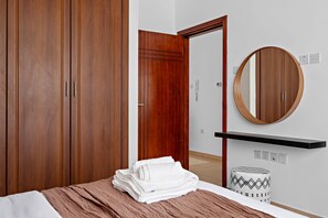 A bedroom with a wooden wardrobe, a round mirror, a bedside table, and a neatly folded white towel. - Silkhaus Near Metro Art XII Tower Pool Gym (Dubai)
