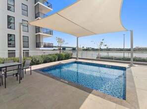 Piscina ao ar livre com borda azulejada, mesa e cadeiras, e um toldo fornecendo sombra.