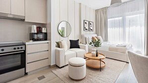 A modern living room with a sofa, ottoman, and coffee table, a kitchen with a stove and microwave, and a large window with sheer curtains. - Silkhaus Canal View Studio Near Ferrari World (Abu Dhabi)