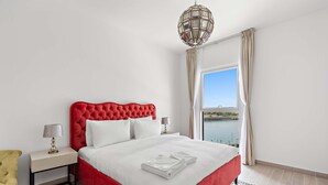 Ein Hotelzimmer mit einem roten, bezogenen Bett, weißen Bettwäsche, einem Nachttisch mit Lampe und Blick auf ein Gewässer durch ein großes Fenster.