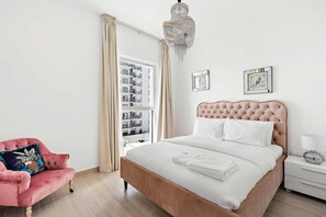 Une chambre à coucher avec un grand lit, un fauteuil rose, un lustre et une fenêtre avec des rideaux.
