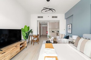A modern living room with a TV, wooden entertainment unit, a sofa, a dining table with chairs, and a kitchen area in the background. - Silkhaus Canalside Cozy Studio Water s Edge (Abu Dhabi)