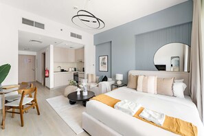Habitación de hotel moderna con una cama grande, un área de comedor con una mesa y sillas, una cocina al fondo y un espejo grande en la pared.