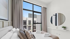 Moderni hotellihuone, jossa on suuri sänky, pyöreä peili ja näkymä kaupunkiin suurista ikkunoista.