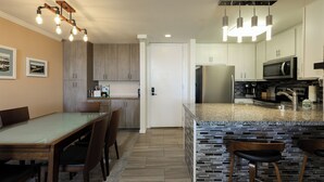 A modern kitchen with granite countertops, stainless steel appliances, and a dining area with a glass-top table and wooden chairs. - Harbor & Ocean View Coastal Retreat – Walk to Beach & Marina (Oceanside)