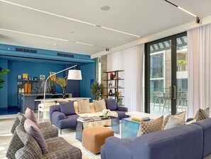 A modern living room with a blue accent wall, a dining area, and a large glass door leading to an outdoor balcony. - Silkhaus Chic Studio Sobha Hartland Kensington (Dubai)