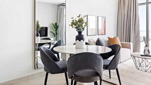 Uma sala de estar moderna com uma mesa de jantar redonda, cadeiras estofadas cinzas, um espelho grande e vista da cidade por grandes janelas.