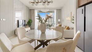 Un salon moderne avec une table ronde en marbre, des fauteuils blancs, une grande plante en pot et une vue sur la ville à travers la porte vitrée.