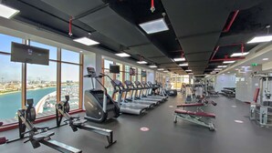 A gym with treadmills, weight machines, and a view of a cityscape through large windows. - Silkhaus Studio on Palm New Modern Sea View (Dubai)
