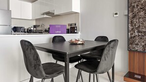 Eine moderne Küche mit weißen Schränken, einem Edelstahlkühlschrank, einer Mikrowelle und einer Kaffeemaschine. Auf der Arbeitsplatte befindet sich ein violettes Schild und auf dem Esstisch eine Fruchtschale.
