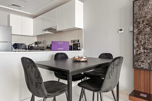 A modern kitchen with white cabinets, a stainless steel refrigerator, a microwave, and a coffee machine. There is a purple sign on the counter and a bowl of fruit on the dining table. - Silkhaus Stylish 2bdr Stay in Radiant Boulevard (Abu Dhabi)