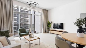 A modern living room with a sofa, armchair, coffee table, and dining table. There is a TV, a plant, and a window with curtains. - Silkhaus Spacious 1BR Najmat Reem Island (Abu Dhabi)