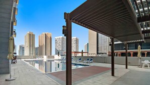 Zona de piscina en la azotea con sillas de descanso y sombrillas, rodeada de edificios altos.