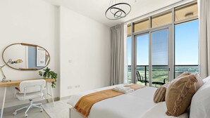 Ein modernes Schlafzimmer mit einem großen Bett, einem Schreibtisch mit Stuhl, einem Spiegel und Blick ins Freie durch große Fenster.