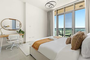 Ein modernes Schlafzimmer mit einem großen Bett, einem Schreibtisch mit Stuhl, einem Spiegel und Blick ins Freie durch große Fenster.