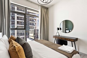 A hotel room with a bed, a desk, a round mirror, and a view of a building through large windows. - Silkhaus Urban 1bdr Near Reem Central Park (Abu Dhabi)