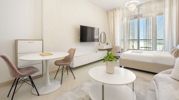 Ein modernes Hotelzimmer mit einer weißen Couch, einem Esstisch, einem Fernseher und einem großen Fenster mit Vorhängen.