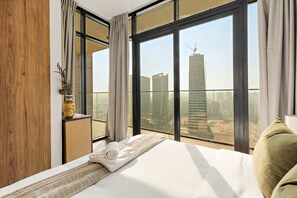 A modern hotel room with a large bed, a wooden panel wall, a vase with dried plants, and a view of a cityscape through large windows. - Silkhaus Luxury 3bdr Stay in Maryah Island (Abu Dhabi)