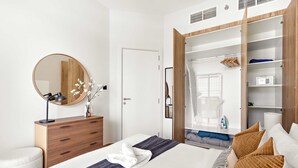 Una habitación de hotel moderna con una cama bien hecha, un aparador de madera, un espejo grande y un vestidor con espacio para colgar ropa.