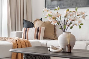 A living room with a sofa, pillows, a vase with flowers, and a table with a candle and a decorative item. - Silkhaus Serene 1bdr Stay Near Reem Mall (Abu Dhabi)