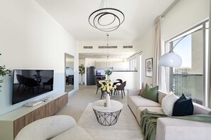 Ein modernes Wohnzimmer mit Flachbildfernseher, rundem Couchtisch und einer Couch. Ein Essbereich mit Tisch und Stühlen ist vorhanden, und im Hintergrund befindet sich eine Küche.