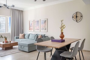 A modern living room with a grey sofa, a wooden dining table, and grey chairs. There are framed pictures on the wall, a vase with flowers, and a clock. - Silkhaus Cozy 1bdr Retreat Dubai Marina (Dubai)