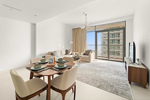 A modern living room with a dining area, large windows, and a view of a high-rise building. - Silkhaus 1bdr w Sofa Bed Private Beach Access (Dubai)