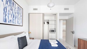 A hotel room with a bed, a blue throw blanket, a white pillow, a framed abstract painting, a wardrobe, and a kitchenette. - Silkhaus Chic Seaview 1bdr Marina Vista Tower (Dubai)
