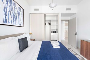 Ett hotellrum med en säng, en blå pläd, en vit kudde, en inramad abstrakt målning, en garderob och ett litet kök.