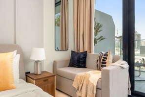 A hotel room with a bed, a sofa, a nightstand with a lamp, a large mirror, and a view of the city through a window. - Silkhaus New Studio Al Maryah Vista Near Reem (Abu Dhabi)