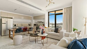 A modern living room with a sofa, ottoman, and coffee table. There is a dining area with a table and chairs. The room has a large window with a view of the city. - Silkhaus Premium Large 3BR Maid Marina Vista (Dubai)