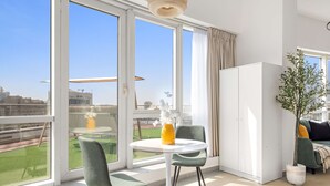 A modern living room with large windows, a round table, two chairs, a sofa, and a potted plant. - Silkhaus Cozy Studio Madison Barsha Heights (Dubai)
