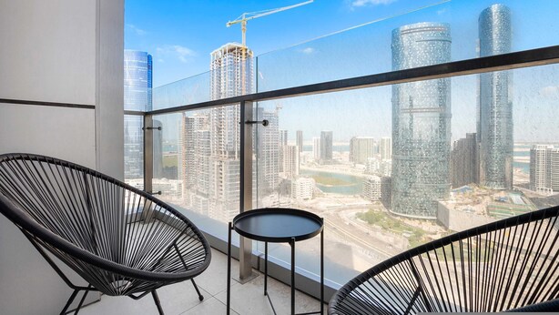 A balcony with two black wire chairs and a small round table, overlooking a cityscape with modern buildings and a construction crane. - Silkhaus Horizon Tower High Floor by Al Fay Park (Abu Dhabi)