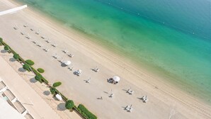 An aerial view of a beach with rows of sun loungers and umbrellas, a clear blue sea, and boats docked in the background. - Silkhaus Emaar Beachfront Tower w Balcony (Dubai)