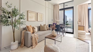 A modern living room with a grey sofa, a coffee table, and a large potted plant. There are two framed artworks on the wall and a balcony with a view. - Silkhaus Elegant 1bdr Retreat Al Maryah Island (Abu Dhabi)