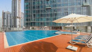 A swimming pool with lounge chairs and umbrellas, surrounded by modern high-rise buildings. - Silkhaus 1bdr in Horizon Tower Al Fay Park Near (Abu Dhabi)
