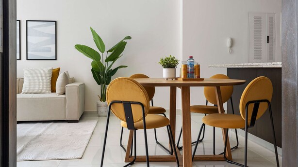 A modern dining area with a round wooden table, six yellow chairs, a potted plant, and a white sofa with yellow cushions. - Silkhaus 1bdr in Horizon Tower Al Fay Park Near (Abu Dhabi)