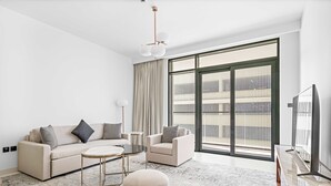 A modern living room with a sofa, armchair, coffee table, and ottoman. There is a large window with curtains, a flat-screen TV, and a ceiling light fixture. - Silkhaus Sea View 1bdr Marina Vista Tower (Dubai)