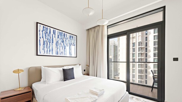 A modern hotel room with a large bed, a framed abstract painting, a nightstand, and a view of a cityscape through a large window. - Silkhaus Cozy 1bdr in Marina Vista Private Beach (Dubai)