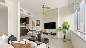 Un salon moderne avec un canapé, une table basse et un téléviseur fixé au mur. On y trouve également un coin repas avec une table et des chaises, ainsi qu’une cuisine à l’arrière-plan.