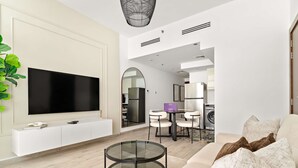 Un soggiorno moderno con una TV a schermo piatto, un tavolino in vetro e un divano con cuscini. C'è una zona pranzo con tavolo e sedie, e una cucina con elettrodomestici sullo sfondo.