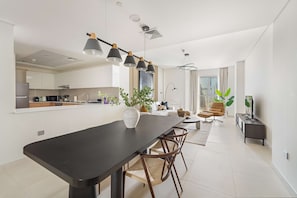 A modern kitchen with white cabinets, a black dining table, and pendant lighting. There is a living area with a sofa and a TV on a stand. - Silkhaus 1bdr in Reem Island in New Modern Tower (Abu Dhabi)