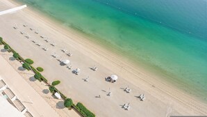 Eine Vogelperspektive zeigt einen Strand mit Reihen von Liegestühlen und Sonnenschirmen, ein klares blaues Meer und eine Marina mit Booten.