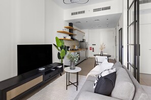 A modern living room with a flat-screen TV, a grey sofa, a small white table, and a kitchen in the background. - Silkhaus Socio Tower Dubai Hills Neighborhood (Dubai)