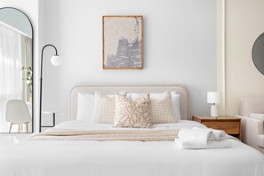Ein ordentlich bezogenes Bett mit weißen Leinentüchern und dekorativen Kissen, ein Nachttisch mit Lampe und ein gerahmtes Kunstwerk an der Wand.