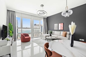 A modern living room with a white sofa, a red armchair, a marble dining table, and a large window offering a view of buildings and a body of water. - Silkhaus Horizon Tower Big Layout Near Park (Abu Dhabi)