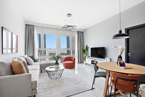A modern living room with a sofa, coffee table, and a dining area with chairs. There is a TV, a door, and large windows with curtains. - Silkhaus Big 1bdr by Al Fay Park Horizon Tower (Abu Dhabi)