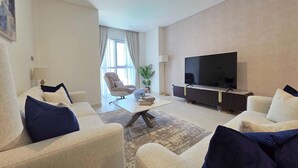 A modern living room with a flat-screen TV, beige sofas with blue cushions, a coffee table, and a large window with curtains. - Silkhaus Serene 1bdr Apartment Al Reem Island (Abu Dhabi)