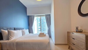 A modern bedroom with a large bed, a wooden dresser, and a view of a building through the window. - Silkhaus Signature 1BR in Reem Island Gate Tower (Abu Dhabi)